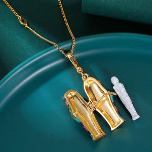 May include: Gold-toned pendant necklace featuring an open, hinged sarcophagus with colorful accents and a small white mummy figure. The necklace is displayed on a teal surface, showcasing the intricate details of the Egyptian-themed jewelry.