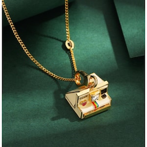 May include: Gold-toned necklace featuring a pendant shaped like a vintage instant camera. The pendant is cream-colored with gold trim, a rainbow stripe, and a small red heart. The chain is a delicate link style.