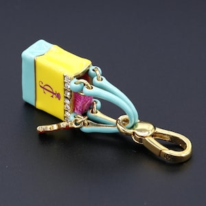 May include: A colorful keychain shaped like a shopping bag. The bag is yellow with a light blue top and bottom, and a pink interior. The keychain has a gold clasp and accents, with a decorative jewel border and a purple logo.