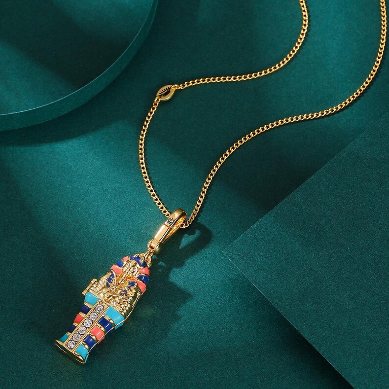 May include: Gold-toned necklace featuring a pendant shaped like an Egyptian sarcophagus. The pendant is adorned with blue, red, and orange enamel, and embellished with clear crystals. The necklace has a delicate chain and a secure clasp, perfect for adding a touch of history to any outfit.