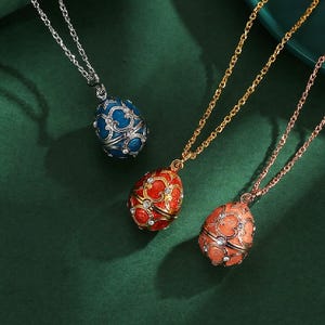 May include: Three egg-shaped pendant necklaces in blue, red, and coral. Each pendant features ornate scrollwork and is accented with small, clear crystals. The necklaces have silver, gold, and rose gold chains.