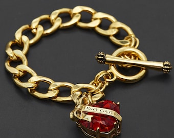 May include: Gold-coloured chain bracelet with a red heart-shaped charm. The charm is embellished with red crystals and the words "Juicy Couture". The bracelet has a toggle clasp.