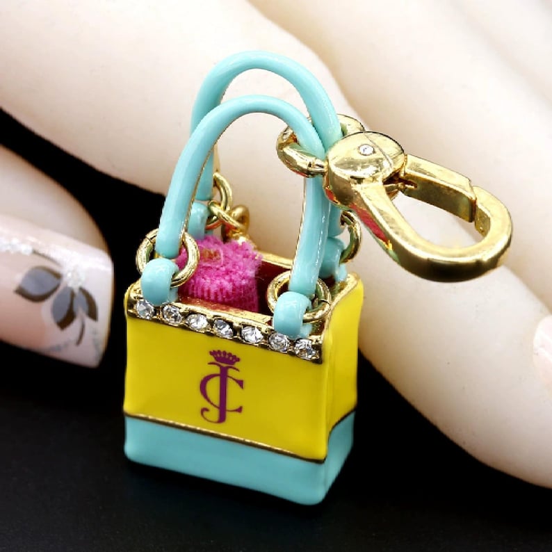 May include: A miniature handbag keychain with a yellow body, turquoise base and handles, and a gold clasp. The bag features a crown and the letter "J" in purple. The top of the bag is lined with small clear crystals.
