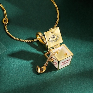 May include: Gold-tone necklace featuring a pendant shaped like a box with the words "Choose Juicy" in red. The box opens to reveal small gold charms. The necklace has a delicate chain and a small charm near the clasp. The Juicy Couture logo is visible.