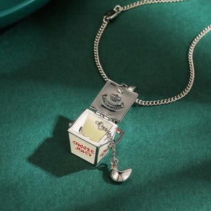 May include: Silver-toned necklace with a pendant shaped like a takeout container. The container is white with red text that reads "CHOOSE JUICY". A silver charm shaped like a crescent moon hangs from the container.