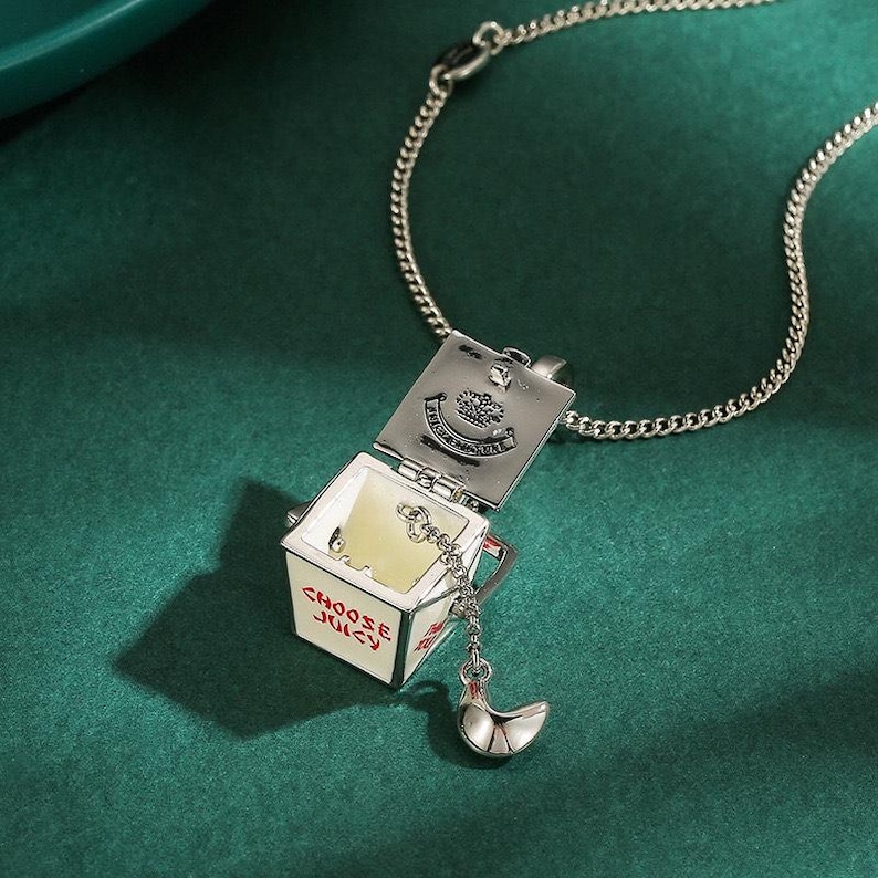 May include: Silver-toned necklace with a pendant shaped like a takeout container. The container is white with red text that reads "CHOOSE JUICY". A silver charm shaped like a crescent moon hangs from the container.