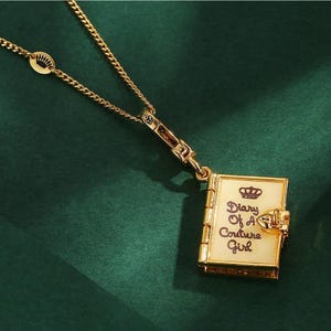 May include: Gold-coloured necklace featuring a book-shaped pendant. The pendant displays the text "Diary Of A Couture Girl" and a crown. The necklace is set against a dark green backdrop.