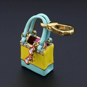 May include: A miniature handbag charm in yellow and turquoise with gold-tone accents. The charm features a pair of red sunglasses and is embellished with rhinestones. The handbag has turquoise handles and a gold clasp.