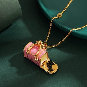 May include: A gold necklace featuring a pink handbag pendant with gold detailing. The handbag opens to reveal a small black dog figurine. The pendant has the text "For Nice Dogs Who Like Stuff".
