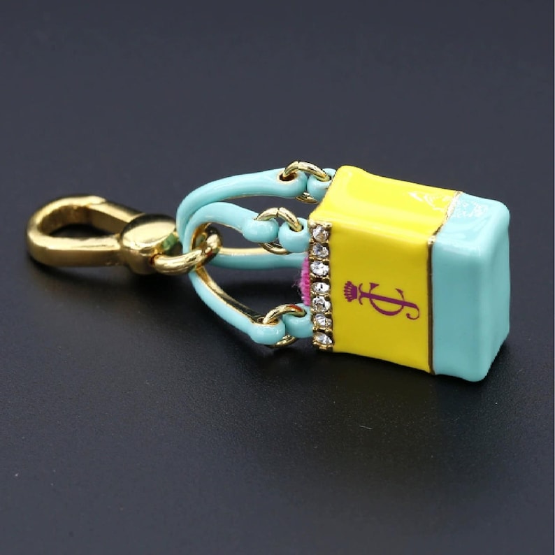 May include: A yellow and turquoise handbag-shaped keychain with gold-tone hardware. The keychain features a small, rectangular bag with a rhinestone-studded border and a purple logo. The handle is turquoise and the clasp is gold.