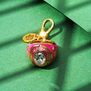 Könnte beinhalten: Ein goldfarbener Schlüsselanhänger mit einem rosa herzförmigen Anhänger. Der Anhänger trägt den Text "SCHOOL COUTURE" in goldfarbenen Buchstaben um einen großen, klaren Kristall. Ein kleines goldenes Etikett mit einer Krone ist angebracht.