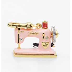 May include: A pink sewing machine charm with gold-tone details. The charm has the word "Juicy" in cursive, a red thread spool, and a heart with a crown. The sewing machine's wheel is decorated with clear crystals.