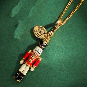 Pode incluir: Colar dourado com um pingente de quebra-nozes em miniatura. O pingente é pintado em vermelho, preto, branco e dourado, com um pequeno bigode e um chapéu preto. Uma etiqueta dourada diz "LIMITED EDITION 2006". O colar está em um fundo verde.