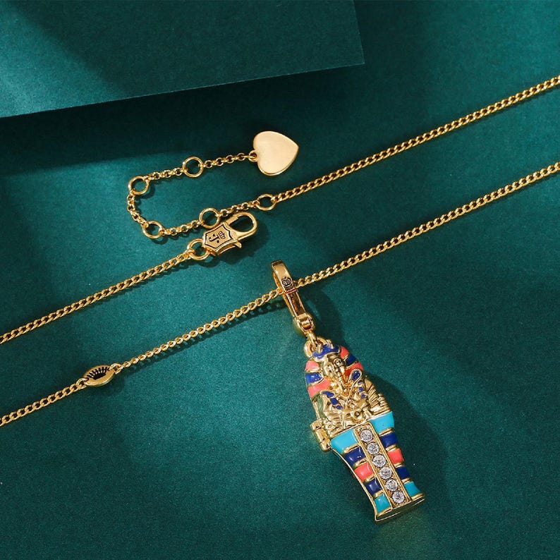 May include: Gold-toned necklace featuring a pendant shaped like an Egyptian sarcophagus. The pendant is adorned with colorful enamel and sparkling gemstones. The necklace has a delicate chain and a heart-shaped charm near the clasp.