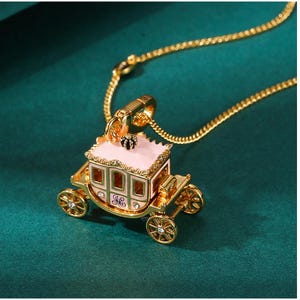 May include: A gold-colored necklace with a pendant shaped like a carriage. The carriage is pink with gold trim and wheels, and a small black crown on top. The necklace is on a dark green background.