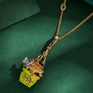 May include: Gold-tone necklace with a pendant featuring a lime green flower pot filled with flowers and a butterfly. The pendant has the words "Juicy Couture" on the front. The necklace is on a dark green background.