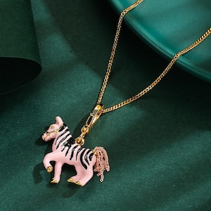 Puede incluir: Un collar de cadena dorada con un colgante en forma de cebra rosa con rayas negras. La cebra tiene una melena y cola rosas. El collar se muestra sobre una superficie verde oscuro.
