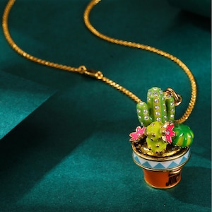 May include: Gold-tone necklace featuring a pendant shaped like a potted cactus. The cactus is green with rhinestone accents, pink flowers, and a green melon. The pot is orange with a blue and white geometric design and the word "JUICY".