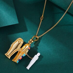 May include: Gold-toned necklace featuring a pendant shaped like an open sarcophagus with colorful accents. A small white mummy figure is attached. The necklace hangs against a dark green background.