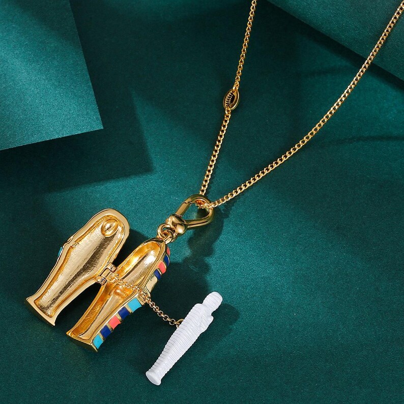 May include: Gold-toned necklace featuring a pendant shaped like an open sarcophagus with colorful accents. A small white mummy figure is attached. The necklace hangs against a dark green background.