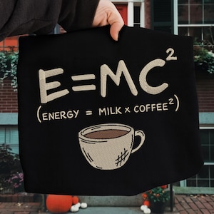 May include: Black tote bag featuring embroidered text and a coffee cup design. The text reads "E=MC² (ENERGY = MILK x COFFEE²)". The coffee cup is beige with a brown interior and a hashtag symbol.