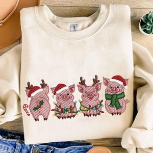 May include: Cream-colored sweatshirt featuring an embroidered design of four pigs dressed for Christmas. Each pig has a Santa hat, and some have antlers, Christmas lights, or a scarf. A candy cane is also included in the design.