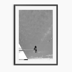 May include: Monochrome framed print featuring a person seated on a structure over water, with birds in flight. The artwork has a minimalist style, emphasizing the silhouette of the figure and the surrounding landscape. The print is framed in black.