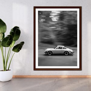 May include: Framed black and white photograph of a silver sports car speeding down a road. The image has a blurred background, suggesting motion. The car is the focal point, set against a backdrop of trees. The frame is dark brown.