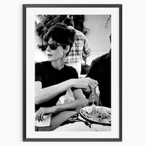 Puede incluir: Impresión enmarcada en blanco y negro de una mujer con gafas de sol, sentada a una mesa, sosteniendo un tenedor sobre un plato de comida. La imagen es un retrato clásico, con una estética vintage.
