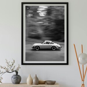 May include: Black and white framed print of a silver sports car speeding down a road. The background is blurred, suggesting motion. The car is the focal point, set against a backdrop of blurred trees. The print is displayed on a white wall.