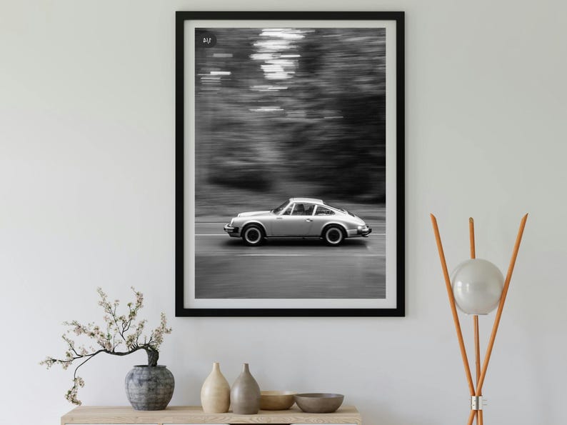 May include: Black and white framed print of a silver sports car speeding down a road. The background is blurred, suggesting motion. The car is the focal point, set against a backdrop of blurred trees. The print is displayed on a white wall.