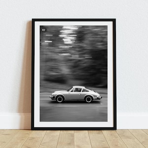 May include: Black and white framed print of a silver sports car speeding down a road. The car is in motion, with a blurred background of trees. The image is set in a black frame and is displayed on a white wall.