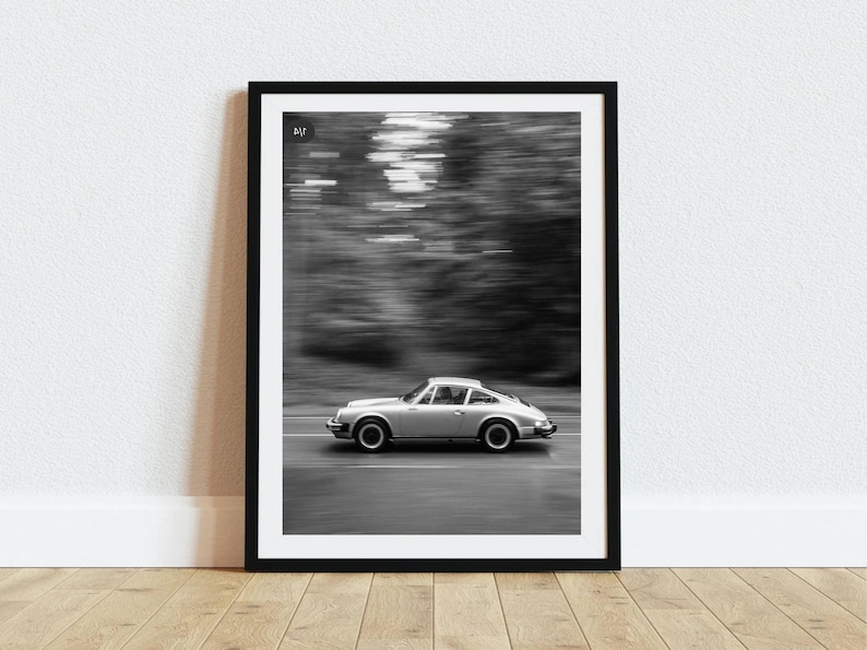 May include: Black and white framed print of a silver sports car speeding down a road. The car is in motion, with a blurred background of trees. The image is set in a black frame and is displayed on a white wall.