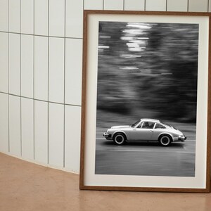 May include: Framed black and white photograph of a silver sports car in motion on a road. The car is a classic model, with a sleek design. The background is blurred, suggesting speed and motion. The frame is a warm brown color.