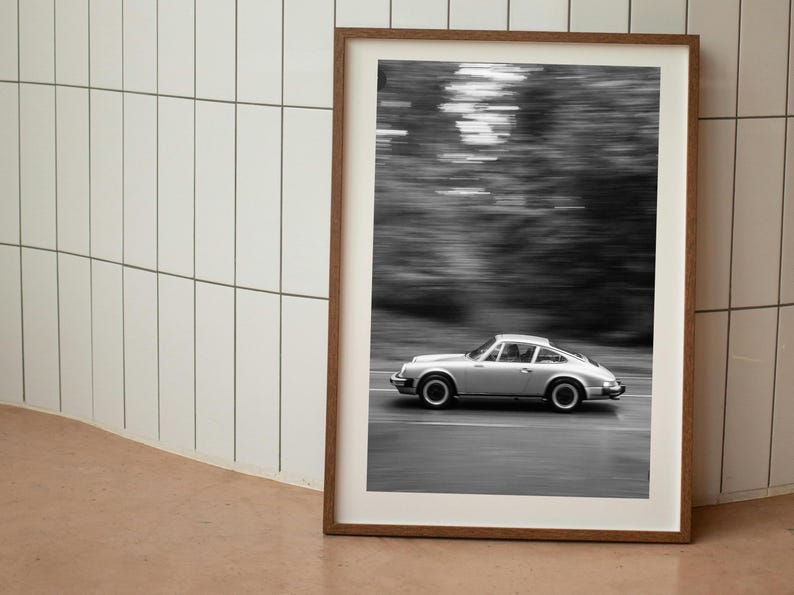 May include: Framed black and white photograph of a silver sports car in motion on a road. The car is a classic model, with a sleek design. The background is blurred, suggesting speed and motion. The frame is a warm brown color.