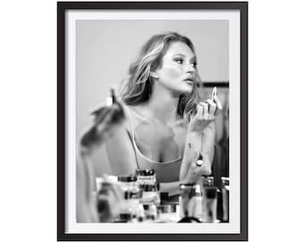 May include: Monochrome framed print showing a woman applying makeup. She holds a lipstick tube, looking into a mirror. Several makeup products are arranged on a table in front of her.