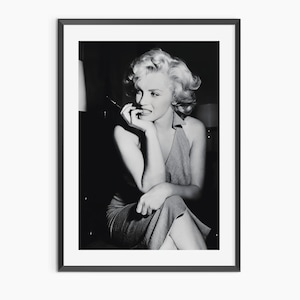 Puede incluir: Impresión enmarcada en blanco y negro que presenta a Marilyn Monroe. La imagen muestra a Monroe sentada, sosteniendo un cigarrillo, con la mano en la barbilla. Lleva un vestido halter y tiene el pelo rubio.
