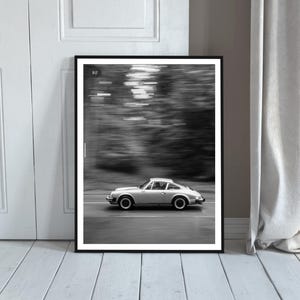 May include: Black and white framed print of a silver sports car in motion on a road. The car is a classic model, with a sleek design. The background is blurred, suggesting speed. The print is displayed against a white door and a light-colored wooden floor.