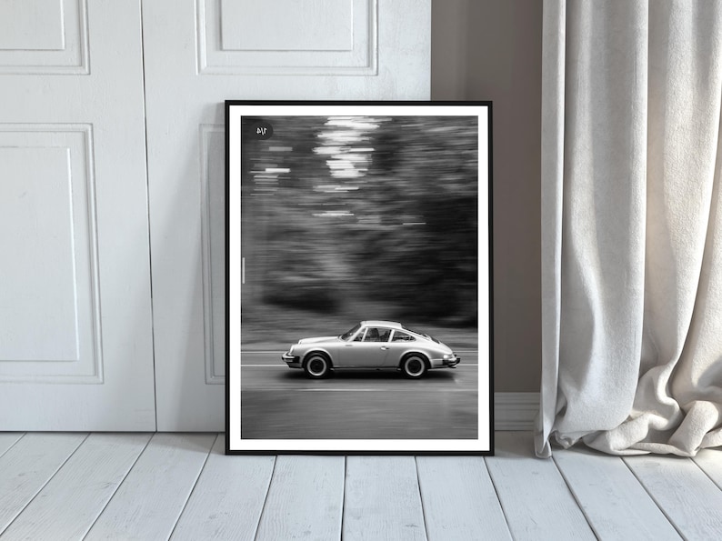 May include: Black and white framed print of a silver sports car in motion on a road. The car is a classic model, with a sleek design. The background is blurred, suggesting speed. The print is displayed against a white door and a light-colored wooden floor.