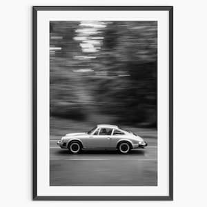 May include: Black and white framed print of a silver sports car in motion. The car is driving on a road with blurred trees in the background, creating a sense of speed and movement. The image is a classic car photograph.
