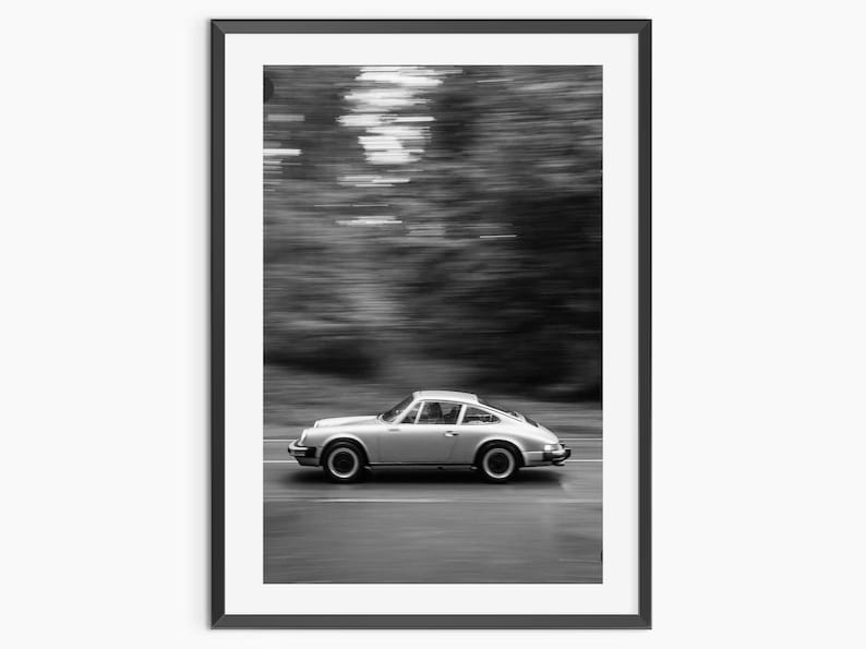 May include: Black and white framed print of a silver sports car in motion. The car is driving on a road with blurred trees in the background, creating a sense of speed and movement. The image is a classic car photograph.