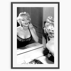 Op de afbeelding: Zwart-wit ingelijste print van een vrouw die zich opmaakt voor een spiegel. De vrouw heeft blond haar en draagt een zwarte top. Een klassiek Hollywood-portret.