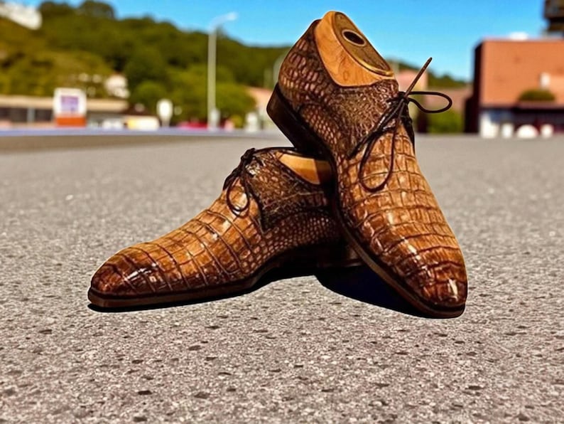 May include: A pair of brown leather dress shoes with a crocodile skin pattern. The shoes have laces and a wooden shoe tree inside. The shoes are on a grey surface with a blurred background of trees and buildings.