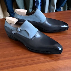 Könnte beinhalten: Zweifarbige Lederschuhe in Schwarz und Hellblau. Die Schuhe haben einen Schnallenverschluss und werden mit hölzernen Schuhspannern gezeigt. Die Schuhe stehen auf einer Holzoberfläche.