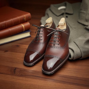May include: A pair of brown leather Oxford shoes with black laces and a polished finish. The shoes have wooden shoe trees inserted. A stack of leather-bound books and a folded olive-green jacket are in the background.