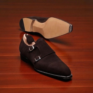 May include: A pair of brown suede double monk strap shoes with silver buckles. The sole of one shoe is visible above the other, showcasing the leather and heel construction. The shoes are displayed on a wooden surface.