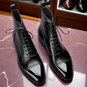 May include: A pair of black leather ankle boots with a glossy finish on the toe caps. The boots have a lace-up design and are displayed on a pinkish-red surface. The boots are approximately 6 inches tall.