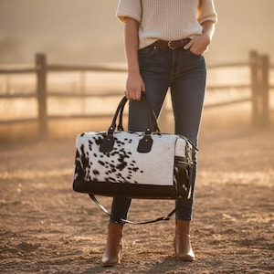 K&ouml;nnte beinhalten: Eine schwarz-wei&szlig;e Kuhfell-Reisetasche mit schwarzen Ledergriffen und -besatz. Die Tasche hat einen abnehmbaren Schultergurt und eine Seitentasche mit Rei&szlig;verschluss. Die Tasche wird von einer Person getragen, die Jeans und braune Stiefel tr&auml;gt.