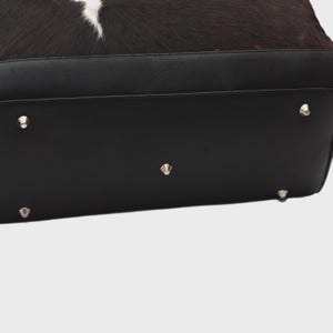 K&ouml;nnte beinhalten: Schwarzer Lederboden einer Handtasche mit silbernen Metallf&uuml;&szlig;en. Der obere Teil der Tasche hat ein Kuhfellmuster in Schwarz, Braun und Wei&szlig;.