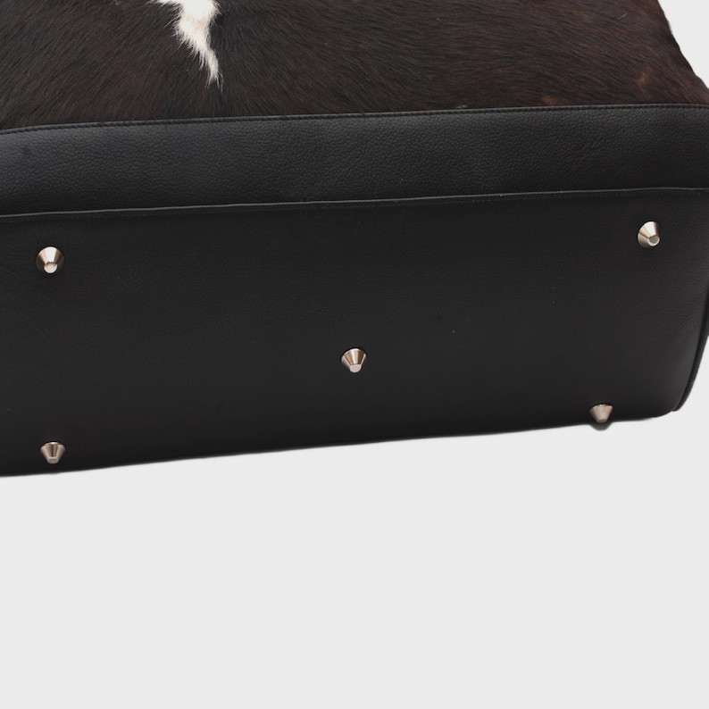 K&ouml;nnte beinhalten: Schwarzer Lederboden einer Handtasche mit silbernen Metallf&uuml;&szlig;en. Der obere Teil der Tasche hat ein Kuhfellmuster in Schwarz, Braun und Wei&szlig;.